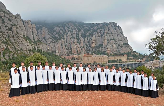 L’Escolania de Montserrat: Secrets de 700 Anys de Música Sagrada 2 Grup de nois de l'Escolania de Montserrat amb hàbits blancs i negres posant davant del monestir de Montserrat i les muntanyes.